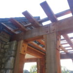 Construction site showing wooden beams and stone pillars under a roof framework.